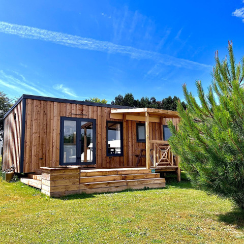 Moderne træhytte i solskin med terrasse og have, set foran Cottage Prestige-lodgen under blå himmel.