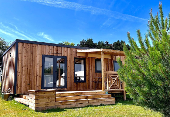 Moderne træhytte i solskin med terrasse og have, set foran Cottage Prestige-lodgen under blå himmel.