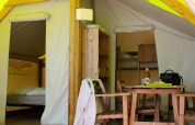 Interior of a safari tent with a dining table, chairs, shelving unit, and visible sleeping areas