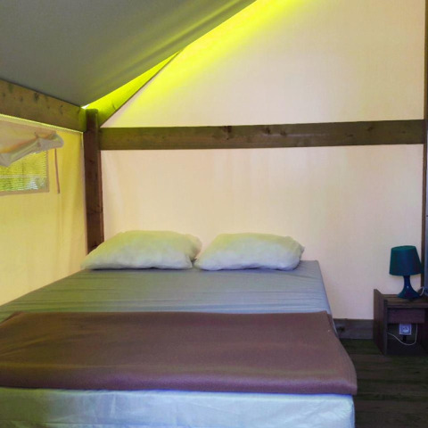 Interior de tienda safari con una cama doble, dos almohadas, mesita de noche y luz ambiental suave.