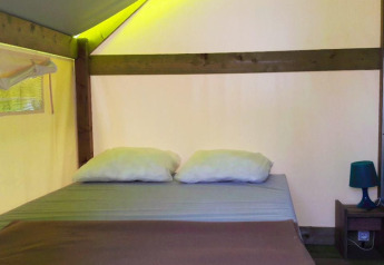 Interior de tienda safari con una cama doble, dos almohadas, mesita de noche y luz ambiental suave.