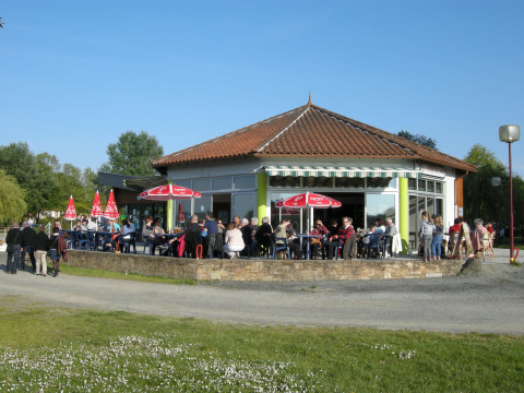 Parque de vacaciones en Pays de la Loire, Francia, con personas sentadas afuera de una cafetería bajo sombrillas rojas.