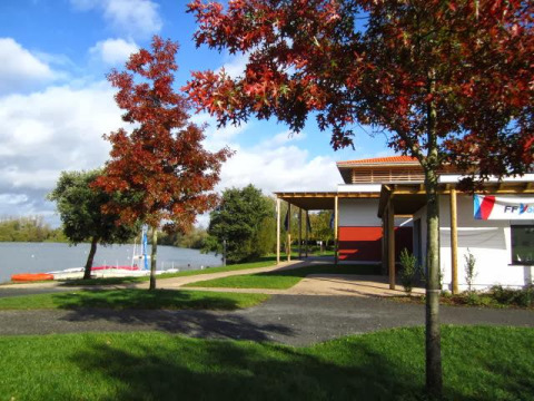 Paisaje otoñal en Camping Seasonova du Chêne, Pays de la Loire, Francia, con lago, edificio y árboles.