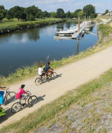 Senda ciclista junto al río en Camping Seasonova du Chêne, Pays de la Loire, Francia, con familias en bicicleta.