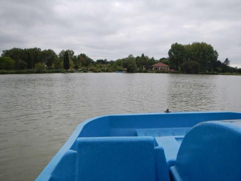Zicht vanuit een blauwe boot op een vijver bij Camping Seasonova du Chêne in Pays de la Loire, Frankrijk, met bomen en paviljoen.