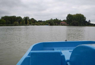 Udsigt fra en blå robåd på en sø ved Camping Seasonova du Chêne i Pays de la Loire, Frankrig, omgivet af træer og en pavillon.