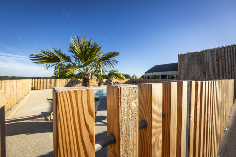 Piscina al aire libre con palmera y valla de madera bajo cielo azul en Domaine Seasonova Saâne et Mer, Normandía.