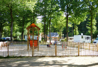Speelplein met houten speeltoestellen en caravans tussen bomen op Camping Seasonova La Forêt, Frankrijk.
