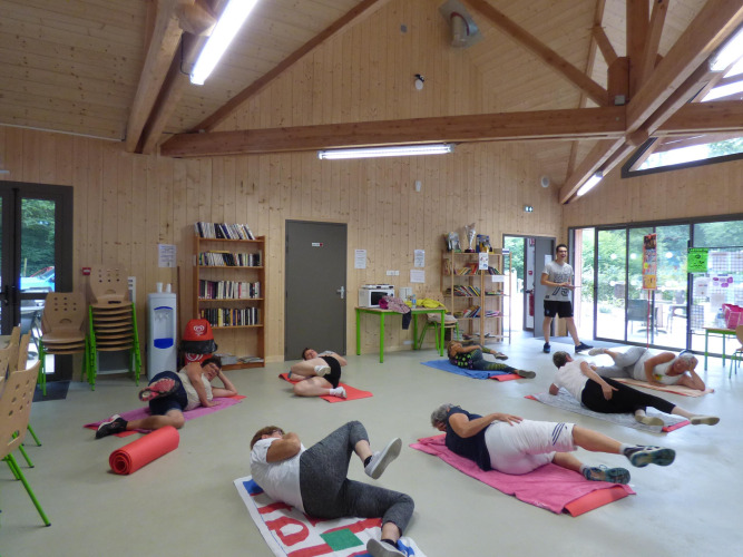 Des personnes participent à un cours de gym en groupe sur des tapis dans une salle en bois clair au Camping Seasonova La Forêt.