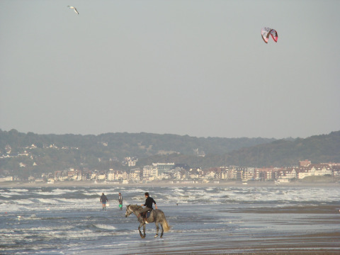 Persona montando a caballo en la playa de Camping Seasonova Le Point du Jour, Normandía, Francia, con ciudad al fondo.