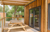 Porche de madera con mesa de picnic junto a un lodge, rodeado de árboles y más cabañas de madera al fondo.