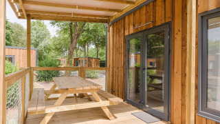 Overdekt houten terras met picknickbank aan een lodge, omgeven door bomen en andere lodges in de buurt.