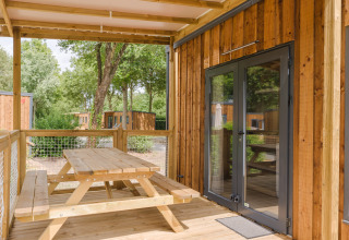 Porche de madera con mesa de picnic junto a un lodge, rodeado de árboles y más cabañas de madera al fondo.
