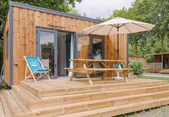 Moderne houten lodge met terras, parasol, picknicktafel en ligstoelen omgeven door groene bomen.