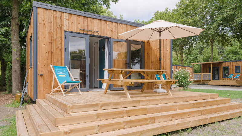 Een modern houten lodge met terras, parasol, picknicktafel en ligstoelen omgeven door groene bomen.