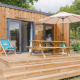 A modern wooden lodge with a terrace, umbrella, picnic table, and deck chairs surrounded by trees.