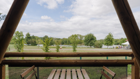 Uitzicht vanop een safaritent-terras op bomen en een meer, met tafel en stoelen, onder een bewolkte hemel.