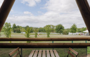 Uitzicht vanop een safaritent-terras op bomen en een meer, met tafel en stoelen, onder een bewolkte hemel.