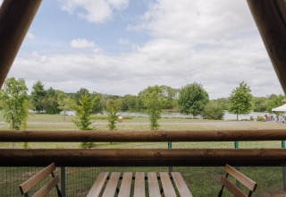Uitzicht vanop een safaritent-terras op bomen en een meer, met tafel en stoelen, onder een bewolkte hemel.