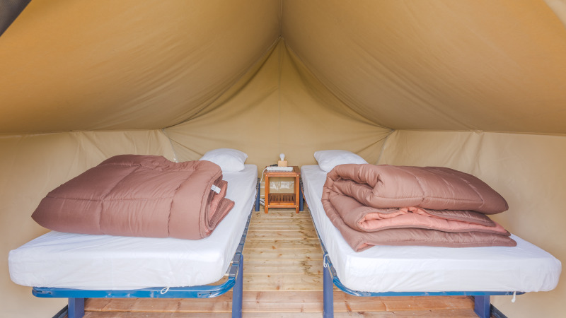 Interior of a safari tent featuring two single beds, brown duvets, and a nightstand on a wooden floor.