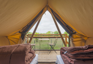 Uitzicht van binnenin een safaritent met bedden, dekens en een houten terras met tafeltje en stoelen.