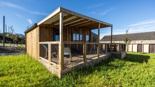 Prestige Cottage lodge met houten buitenkant, overdekt terras en grasveld onder een heldere blauwe lucht.