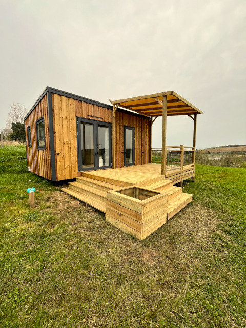 Cabaña llamada Cottage con terraza cubierta, escalones de madera y rodeada de césped y cielo nublado.