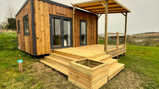 Cabaña llamada Cottage con terraza cubierta, escalones de madera y rodeada de césped y cielo nublado.