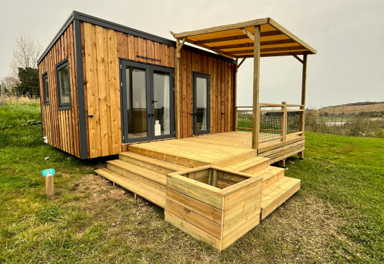 Træhytte ved navn Cottage med overdækket terrasse og trappe, omgivet af grønt græs og åben himmel.