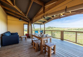 Terraza cubierta de una tienda safari Ecolodge con mesa de comedor, sofá y vistas al campo abierto.
