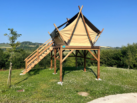Tienda safari elevada sobre campo de césped, con escaleras de madera y cielo azul despejado.