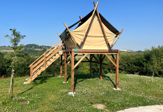 Elevated safari tent on a grassy field with wooden stairs, surrounded by greenery and blue sky.