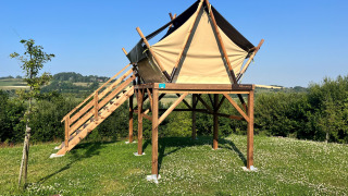 Tienda safari elevada sobre campo de césped, con escaleras de madera y cielo azul despejado.