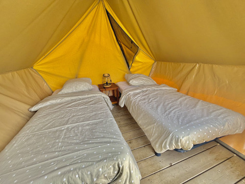 Two single beds with bedding inside a spacious glamping tent on wooden floor, lantern on nightstand.