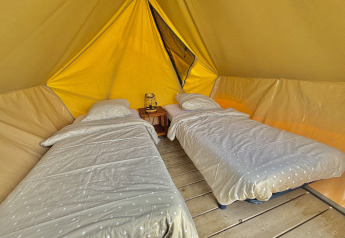 To enkeltsenge med sengetøj i et rummeligt glamping-telt med trægulv og lanterne på natbordet.