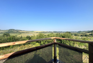 View from a safari tent deck with wooden railing overlooking green fields, hills, and a clear blue sky.