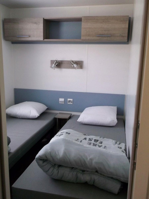 Zwei Einzelbetten mit grauer Bettwäsche in einem kompakten Zimmer im Cottage Zen, Camping Seasonova La Forêt, Frankreich.