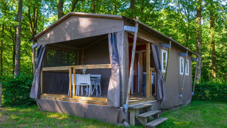 Tienda safari llamada Mobilodge ubicada en un bosque verde con terraza y mesa y sillas afuera.