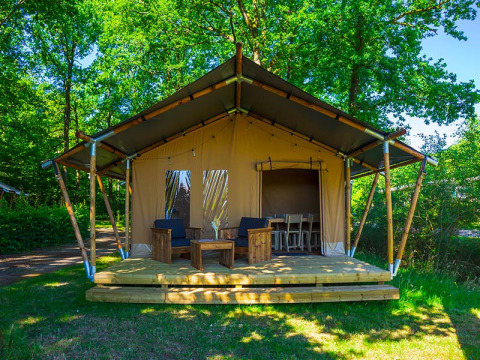 Safari-Zelt Slow Lodge mit Holzterrasse und Sitzgelegenheit im Freien, umgeben von grünen Bäumen.