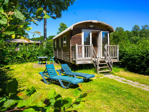 Gipsy wagon cabin with deck chairs and table in a sunny green garden, surrounded by lush trees.
