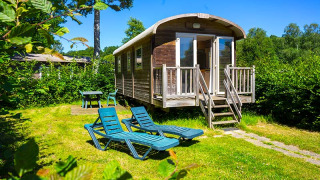 Gipsy wagon cabin with deck chairs and table in a sunny green garden, surrounded by lush trees.