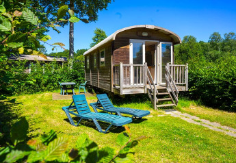 Gipsy wagon cabin with deck chairs and table in a sunny green garden, surrounded by lush trees.