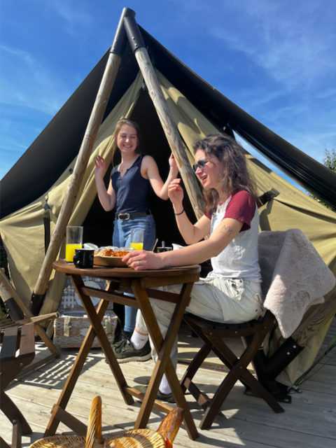 Dos mujeres desayunan al aire libre frente a una tienda tipi, sonrientes bajo el cielo azul y soleado.
