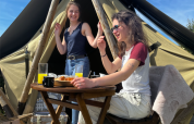 Dos mujeres desayunan al aire libre frente a una tienda tipi, sonrientes bajo el cielo azul y soleado.