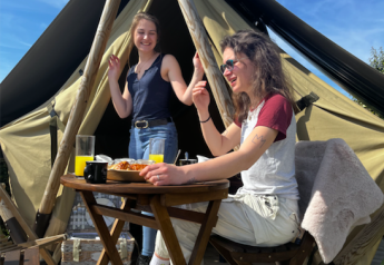 Dos mujeres desayunan al aire libre frente a una tienda tipi, sonrientes bajo el cielo azul y soleado.