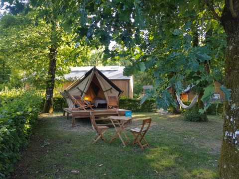 A cozy teepee tent on a wooden deck in a leafy camping area, with outdoor chairs and a hammock.