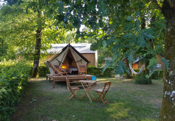 A cozy teepee tent on a wooden deck in a leafy camping area, with outdoor chairs and a hammock.
