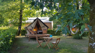 A cozy teepee tent on a wooden deck in a leafy camping area, with outdoor chairs and a hammock.