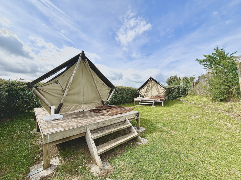 Twee glamping tenten op houten terrassen in een grasrijke buitenruimte met een blauwe, bewolkte lucht.