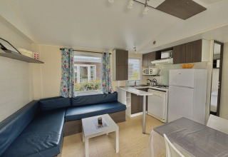 Modern glamping accommodation with blue L-shaped sofa, compact kitchen, and window with colorful curtains.